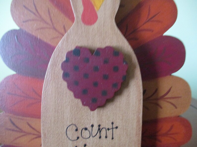 Op de afbeelding: Houten kalkoenfiguur met een rood hart met zwarte stippen en het woord "Count" eronder. De kalkoen heeft bruine, oranje en rode veren.