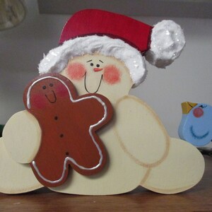 Puede incluir: Una figurita de mu&ntilde;eco de nieve de madera con un gorro de Pap&aacute; Noel rojo y blanco, sosteniendo una galleta de jengibre y una figurita de p&aacute;jaro azul.