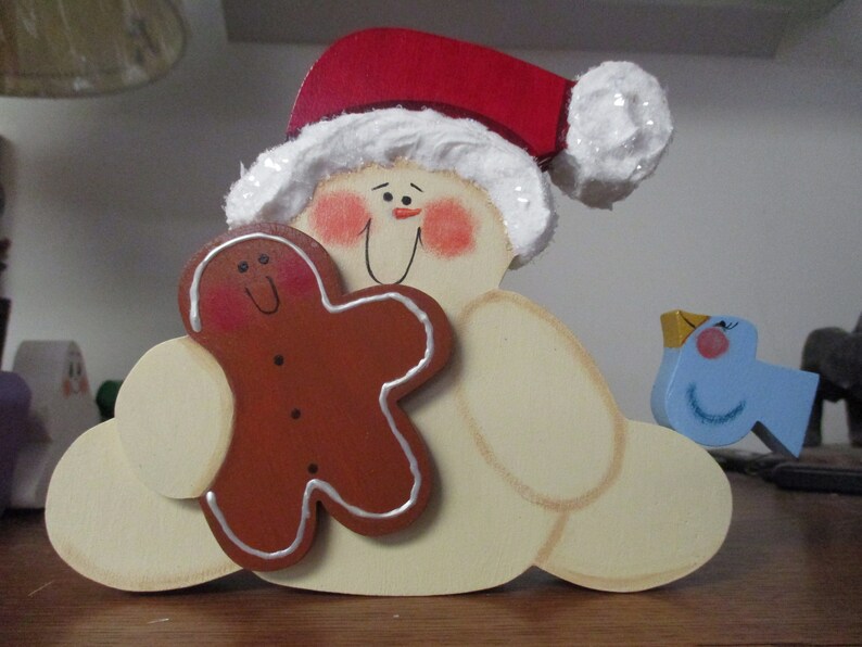 Puede incluir: Una figurita de mu&ntilde;eco de nieve de madera con un gorro de Pap&aacute; Noel rojo y blanco, sosteniendo una galleta de jengibre y una figurita de p&aacute;jaro azul.