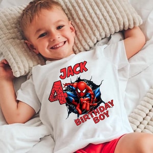 May include: White t-shirt featuring a graphic of Spider-Man bursting through a wall, with the text "JACK 4 BIRTHDAY BOY". The number 4 is designed with a spiderweb. The shirt is worn by a child.