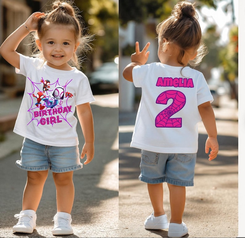 May include: A white t-shirt with a Spider-Man graphic and the text "BIRTHDAY GIRL" in pink. The back of the shirt has the name "AMELIA" and the number "2" in pink. The outfit includes denim shorts and white trainers.