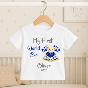 Könnte beinhalten: Weißes T-Shirt mit dem Text "My First World Cup" in Blau und Grau. Ein Teddybär mit blauem Fußballtrikot und schottischen Flaggen ist abgebildet. Der Name "Oliver" und das Jahr "2026" sind ebenfalls auf dem Shirt aufgedruckt.