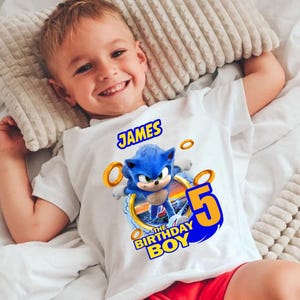 Könnte beinhalten: Weißes T-Shirt mit einem Sonic the Hedgehog-Motiv, goldenen Ringen und dem Text "JAMES", "5" und "THE BIRTHDAY BOY". Das T-Shirt wird von einem Kind getragen.