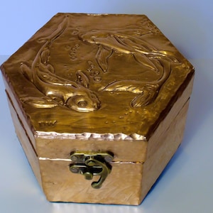 Puede incluir: Una caja hexagonal dorada con tapa con bisagras y cierre. La tapa presenta un relieve de dos peces koi nadando entre burbujas. La caja tiene una superficie texturizada y un acabado metálico, lo que sugiere un objeto decorativo.