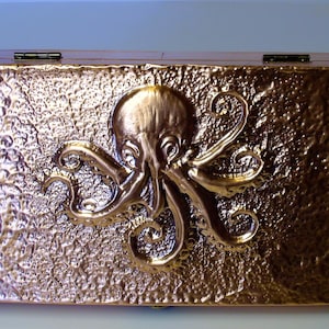 Puede incluir: Una caja rectangular de color bronce con una superficie texturizada y una tapa con bisagras. Un pulpo dorado detallado está fijado en la parte delantera. La caja tiene un brillo metálico y probablemente se utiliza para el almacenamiento.