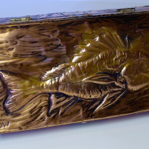 Puede incluir: Una caja rectangular dorada metálica con tapa con bisagras. La caja presenta un relieve detallado de un pez en la parte delantera. La caja tiene una superficie texturizada y probablemente está hecha de metal.