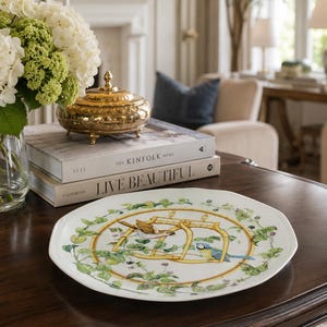May include: A white porcelain plate with a scalloped edge featuring a central illustration of two birds perched on a bamboo-style frame, surrounded by a decorative border of green leaves, vines, and small berries painted in a detailed style.