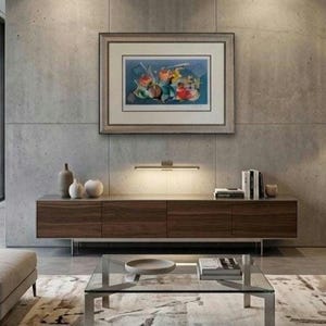 May include: A rectangular framed abstract painting featuring vibrant blue, red, and yellow shapes hangs on a concrete wall above a long, low-profile wooden media console with four cabinet doors and a slim, horizontal LED light fixture mounted below.