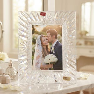 May include: A rectangular clear crystal picture frame featuring a deep-cut sunburst pattern along the border. The frame contains a photograph of a couple in wedding attire and includes a small red label at the top and a gold label at the bottom.