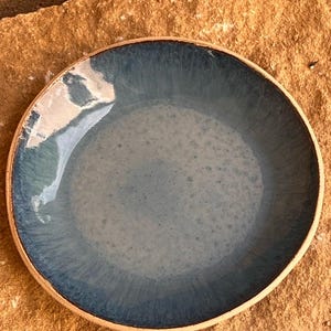 Op de afbeelding: Een ronde keramische schaal met een blauw glazuur en een natuurlijke, ongeglazuurde rand. De binnenkant van de schaal heeft een verloop van blauwtinten, van een lichter midden naar een donkerdere rand. De schaal is waarschijnlijk handgemaakt.