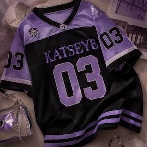 Op de afbeelding: Een zwart en lavendelkleurig V-hals shirt met de tekst KATSEYE in paarse letters op de borst. Het kledingstuk toont het nummer 03 in grote paarse cijfers op de voorzijde en zwarte cijfers op de mouwen, afgewerkt met paarse en zwarte gestreepte boorden.