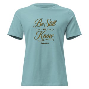 May include: A light blue short-sleeved t-shirt featuring the brown text "Be Still and Know" in a cursive script font, with the smaller text "Psalm 46:10" printed directly underneath in a simple, clean, sans-serif brown typeface.