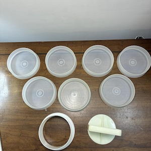 May include: A collection of seven circular, translucent white plastic lids with raised rims, accompanied by one separate white plastic ring and one circular white plastic press tool featuring a central handle for manual operation.