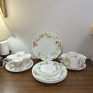 Puede incluir: Un juego de vajilla de cerámica blanca con platos y tazas decorados con un colorido patrón de frutas que incluye peras, manzanas y bayas. La colección incluye una fuente grande, una pila de platos pequeños y varias tazas con platillos.