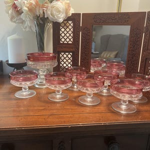 Könnte beinhalten: Ein Set aus klaren Glasdessertschalen und einer passenden Fußschale, jeweils mit einem breiten, cranberryroten Rand und einem strukturierten Pressglasboden. Das Set besteht aus sieben einzelnen Fußschalen und einer höheren Fußschale.