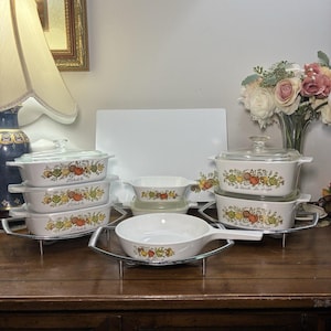 May include: A collection of white ceramic bakeware pieces featuring a colorful vegetable pattern and clear glass lids. The set includes rectangular dishes, a small bowl, and a handled skillet, all resting on silver metal wire stands.