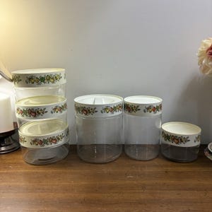 May include: A set of clear glass canisters with off-white lids, featuring a decorative band of colourful fruit illustrations including apples, pears, and grapes. The collection includes one stackable three-tier unit and three individual canisters.