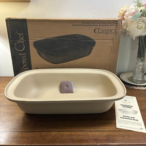 May include: A beige rectangular stoneware bowl with a small purple scraper inside. The bowl is positioned in front of its original cardboard packaging labelled The Pampered Chef Classics Collection and includes a printed recipe and instruction book.