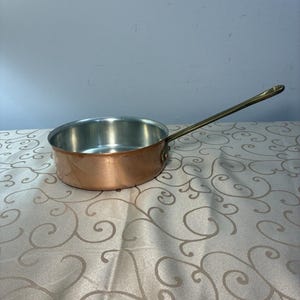 May include: A round copper sauté pan with a polished metallic interior and a long, slender brass handle attached to the side with two rivets. The exterior of the pan features a warm, reddish-brown copper finish with a smooth, reflective surface.