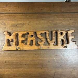 Puede incluir: Un perchero de pared horizontal de madera con la palabra MEASURE recortada en una fuente estilizada. La veta de la madera de color marrón claro natural es visible en toda la pieza. Tres pequeños ganchos de metal están fijados al borde inferior.