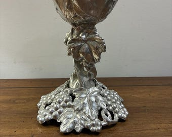 May include: A silver-colored metal candle holder featuring a wide, flared top rim and a central stem decorated with sculpted grape leaves. The base is circular and heavily detailed with clusters of grapes and textured foliage in a metallic finish.