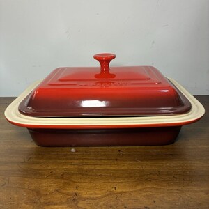Pode incluir: Uma assadeira retangular de ferro fundido esmaltado Le Creuset com tampa a condizer. O exterior apresenta um gradiente de vermelho cereja profundo na base para um vermelho alaranjado brilhante na tampa, com rebordo e puxador creme.