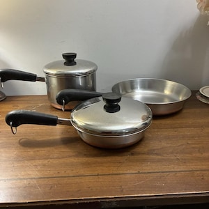 Puede incluir: Un juego de tres piezas de cocina de acero inoxidable con bases de color cobre y mangos negros. El conjunto incluye un cazo profundo con tapa, una sartén poco profunda con tapa a juego y una sartén abierta, todas con superficies de metal pulido.