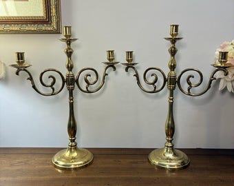 May include: A pair of polished brass candelabras, each featuring a central candle holder flanked by two curved arms with additional candle holders. The metal stands have a smooth, reflective finish with circular bases and scrollwork.