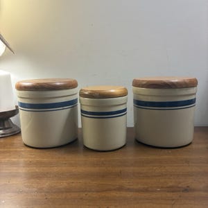 May include: Three cylindrical off-white ceramic canisters of varying sizes, each featuring two horizontal navy blue stripes around the body and a circular wooden lid. The canisters are arranged in a row, with the smallest one positioned in the center.