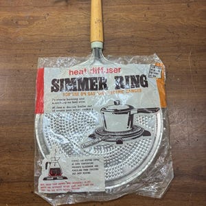 May include: A metal heat diffuser simmer ring with a light wood handle, enclosed in original clear plastic packaging. The packaging features text including 'heat diffuser', 'SIMMER RING', and instructions for use on gas and electric ranges.