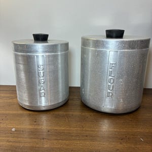 May include: Two cylindrical brushed aluminum canisters with black knobs on the lids. The smaller canister has the word SUGAR embossed vertically on the front, and the larger canister has the word FLOUR embossed vertically on the front.