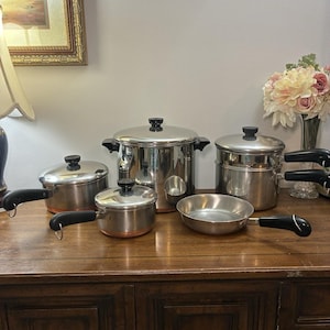 Puede incluir: Un juego de utensilios de cocina de acero inoxidable que incluye una olla grande, dos cacerolas con tapa, un inserto para baño maría y una sartén. Cada pieza tiene un acabado plateado pulido, bases con tonos cobrizos y mangos negros.