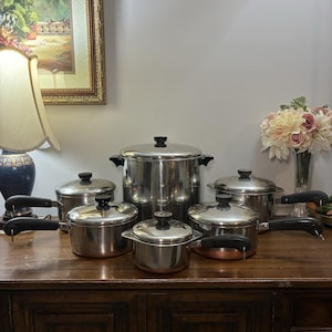 May include: A set of stainless steel cookware featuring a large stockpot and several smaller pots and pans with lids. Each piece has a polished silver finish, black handles, and a visible copper-colored base, arranged in a group on a wooden surface.