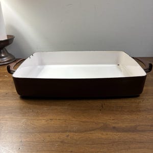 May include: A rectangular baking dish with a dark brown exterior and a smooth white interior. The dish features two curved handles on opposite sides and shows minor signs of wear along the top rim, including small chips in the dark brown finish.