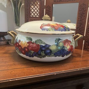 Puede incluir: Una olla redonda de metal esmaltado blanco con tapa a juego, que presenta un colorido patrón pintado de varias frutas, incluyendo manzanas, peras y bayas. La olla tiene dos asas laterales doradas y un pomo dorado en el centro de la tapa.