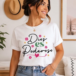 May include: White t-shirt with the text "Dios es Poderoso" in black script, decorated with pink, green, and yellow floral and star accents. The shirt is paired with blue jeans.