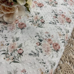 Può includere: Tessuto bianco con un delicato motivo floreale con rose rosa e pesca, foglie verdi e piccoli pois bianchi. Il tessuto è piegato, mostrando il design dettagliato. Un bouquet di fiori è nell'angolo in alto a sinistra.