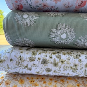 May include: A stack of four folded rolls of ribbed fabric. The top roll features a pink and white floral pattern. The second roll is sage green with white daisy prints. The third roll is white with small green floral sprigs. The bottom roll is yellow floral.