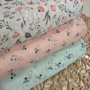 May include: Three rolls of floral fabric in white, pink, and mint green. The white fabric features pink flowers and gray leaves. The pink and mint green fabrics have small, dark floral patterns. The fabrics are stacked on a beige crocheted surface.