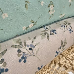 May include: Two rolls of fabric with a textured grid pattern. The top roll is light mint green with a delicate white floral print. The bottom roll is light gray with a botanical print featuring dark blue berries and muted green leaves.