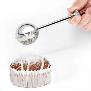 Könnte beinhalten: Ein Puderzuckersieb aus Edelstahl wird über einem Schokoladen-Cupcake in einer weißen Papierform gehalten. Das Sieb hat einen runden, perforierten Kopf und einen langen Griff. Der Cupcake ist mit Puderzucker bestäubt.