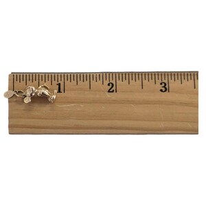 Pode incluir: Um berloque dourado em forma de rato de desenho animado, com aproximadamente 2,5 cm de comprimento, com uma pequena etiqueta anexada. O berloque est&aacute; sobre uma r&eacute;gua de madeira.