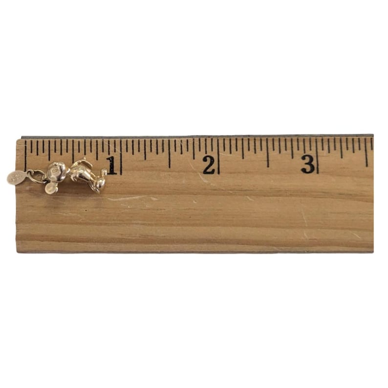Pode incluir: Um berloque dourado em forma de rato de desenho animado, com aproximadamente 2,5 cm de comprimento, com uma pequena etiqueta anexada. O berloque est&aacute; sobre uma r&eacute;gua de madeira.
