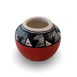 May include: A small, round, terracotta-colored ceramic pot with a wide opening. The upper portion features a black band decorated with white geometric line patterns and small red triangular accents, while the rim is finished in white.