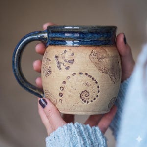 Könnte beinhalten: Ein Keramikbecher mit beigefarbenem, texturiertem Körper, der mit geprägten fossilartigen Mustern wie einer Spiralschale und Blattabdrücken versehen ist. Der Rand und der Henkel sind mit einer dunkelblauen Glasur versehen, die einen Kontrast bildet.