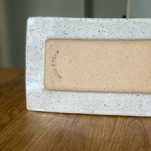 May include: A rectangular ceramic tray with a speckled white border and a tan interior. The words "Julia & Dean" are stamped on the side. The tray is on a wooden surface.