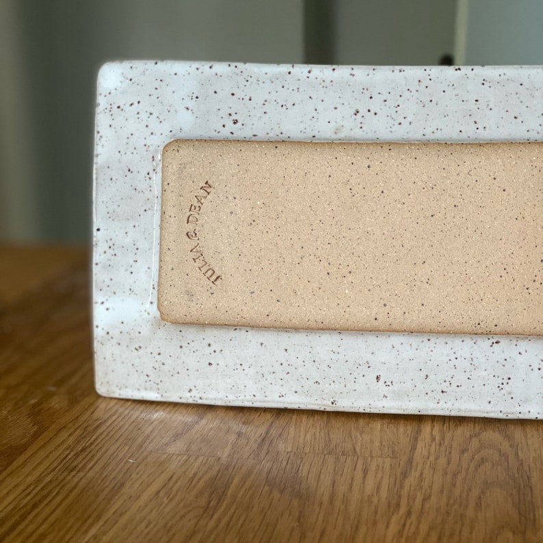 May include: A rectangular ceramic tray with a speckled white border and a tan interior. The words "Julia & Dean" are stamped on the side. The tray is on a wooden surface.