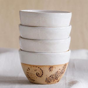 May include: A stack of four white ceramic bowls. The top three bowls are plain white. The bottom bowl has a tan base with a decorative pattern of sea shells and other sea life. The bowls are stacked on top of each other.