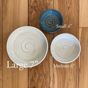 Może przedstawiać: Trzy ręcznie robione ceramiczne miski o różnych rozmiarach. Duża miska ma średnicę 17,78 cm, średnia miska 15,24 cm, a mała miska 10,16 cm. Duże i średnie miski są w kolorze ecru, a mała miska jest niebieska.