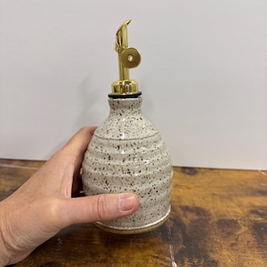 Pode incluir: Um dispensador de óleo de cerâmica com esmalte creme salpicado e textura canelada horizontal. A garrafa possui um bico de metal dourado com mecanismo de tampa articulada, oferecendo um design funcional para uso na cozinha e armazenamento.
